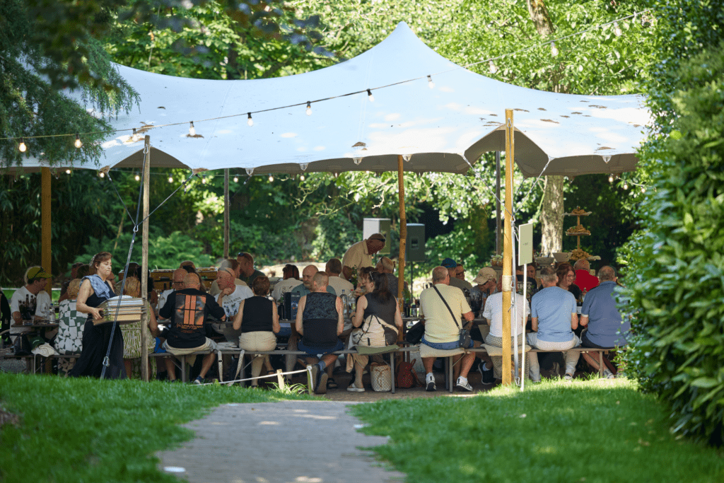 A group of people sit at long tables under a large white marquee outdoors, surrounded by greenery and fairy lights, enjoying shared experiences at a lively social gathering on a sunny day.