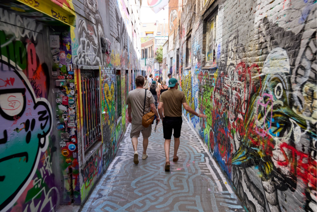 Two people share a walk down a narrow alley covered in colourful graffiti and street art, where vibrant designs, stickers, and murals celebrate shared experiences. Other people can be seen further ahead in the alley.