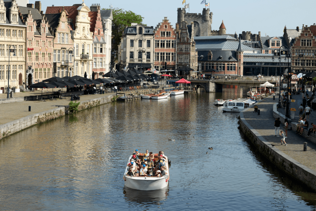 A boat with tourists sails down a canal lined with historic, ornate buildings and outdoor cafés under umbrellas in a lively European city, where shared experiences bring people together along the bustling waterfront promenade.