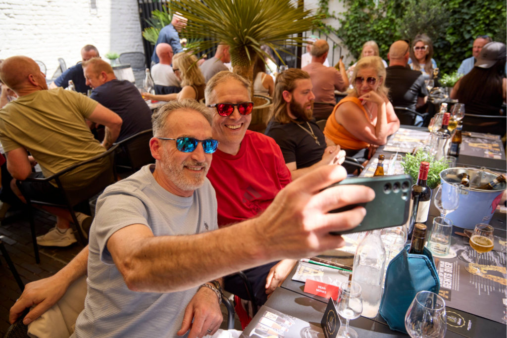 Two smiling men wearing sunglasses take a selfie at an outdoor restaurant table, capturing shared experiences with friends enjoying food and drinks on a sunny day. The lively, relaxed scene is framed by plants in the background.