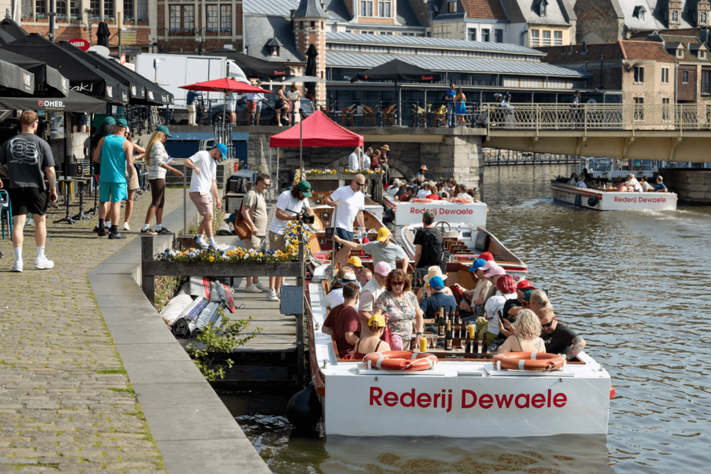 People are enjoying drinks and sharing experiences on boats labelled Rederij Dewaele, moored along a canal in a lively European city, with buildings, cafés, and a bridge in the background.