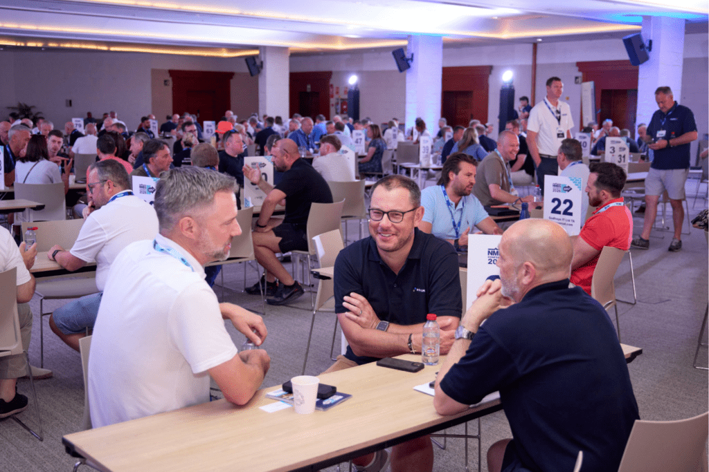A large group of people sit at tables in a spacious, well-lit room with numbered signs, engaging in conversation during a networking or business event. A Bespoke Exhibition Stand is visible, enhancing the professional atmosphere.
