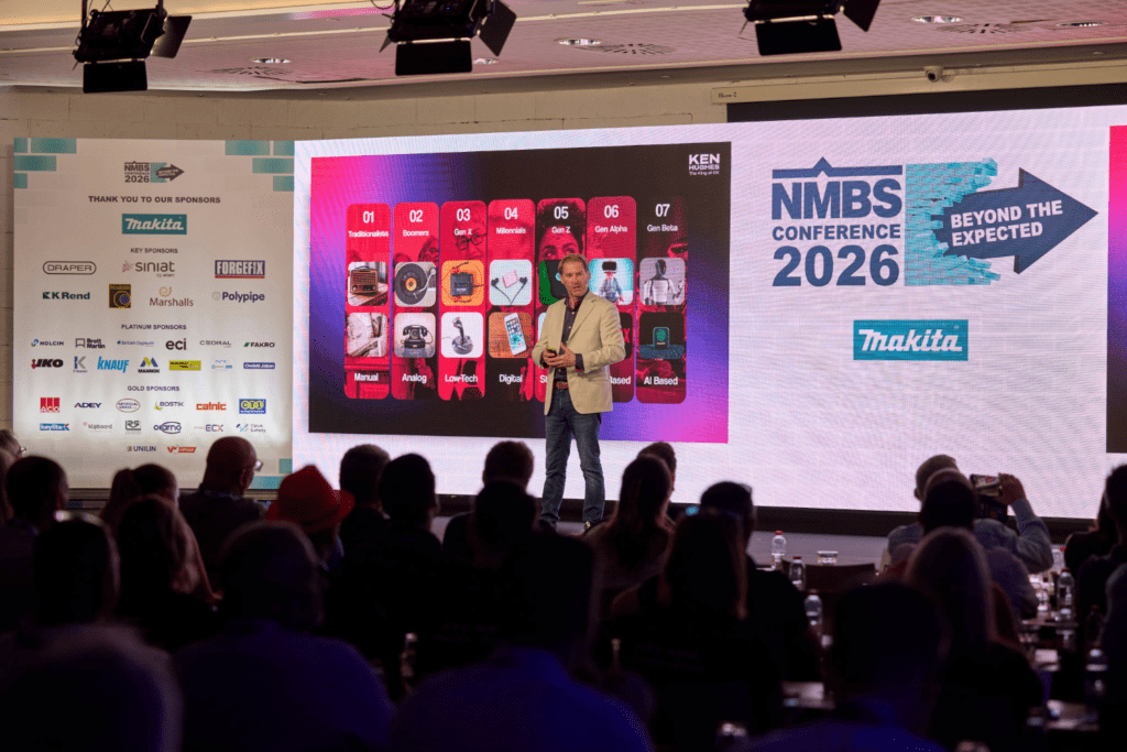 A speaker stands on stage at the NMBS Conference 2026, presenting in front of a large screen. The audience watches attentively, surrounded by conference banners, sponsor logos, and a striking bespoke exhibition stand.