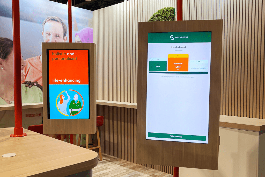 Two digital screens: one displays holistic and personalised, life-enhancing illustrations; the other shows a leaderboard with scores, fostering shared experiences. The setup is in a modern, light-wood booth with red accents.