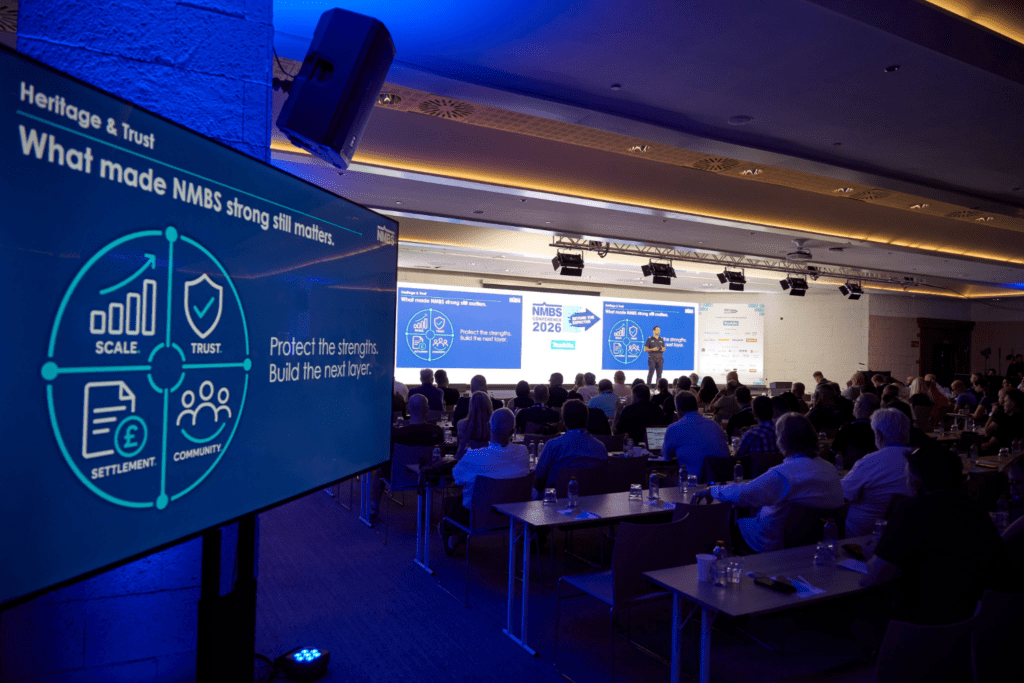 A speaker presents to an audience in a conference room with blue lighting beside a bespoke exhibition stand. A large screen displays a diagram with icons and the words: Scale, Trust, Settlement, Community, and Protect the strengths. Build the next layer.