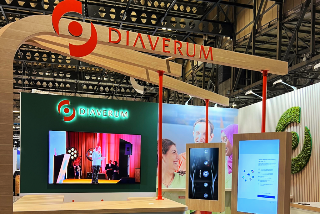 A trade show stand for Diaverum features a large logo, a screen with a presentation, an interactive touchscreen display, and a backdrop with photos of smiling people and green leaf designs, highlighting the power of Shared Experiences.