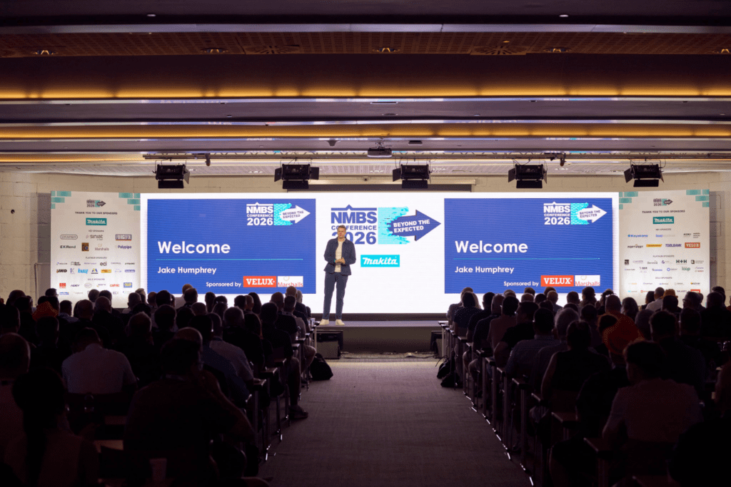 A speaker stands on stage in front of a large screen displaying NMBS Conference 2026 and Welcome to a seated audience in a dimly lit hall, with sponsor logos and a striking Bespoke Exhibition Stand visible on the screen.
