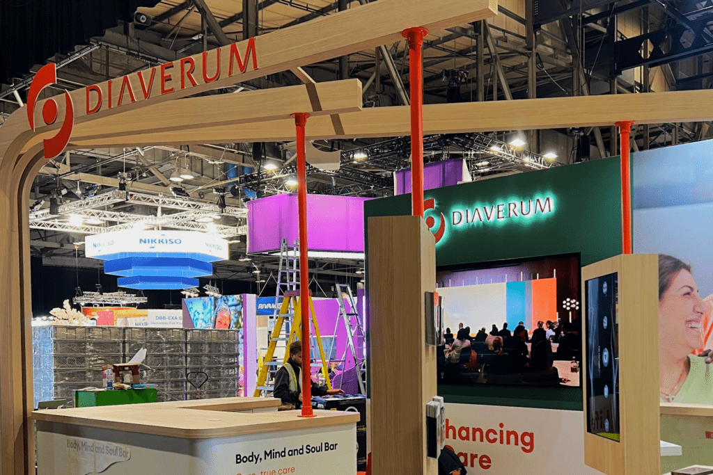 A trade show stand for Diaverum features a curved wooden arch, bright lighting, a large screen displaying shared experiences among people, and colourful structures in the background. A ladder and setup materials are also visible.