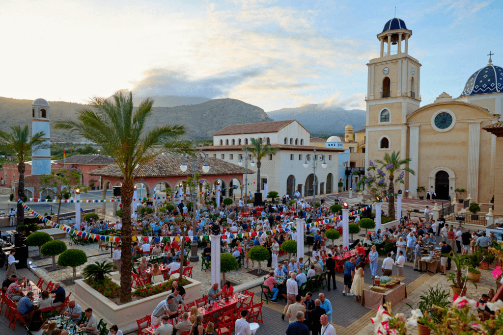 A lively outdoor festival fills a town square with people dining at colourful tables and festive banners overhead, while a bespoke exhibition stand adds flair amid historic Mediterranean-style buildings and a church, all set against a mountainous sunset backdrop.