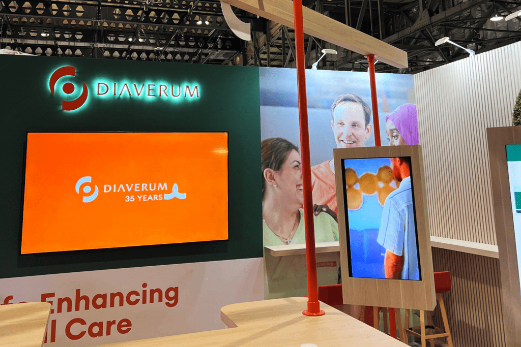 A Diaverum exhibition stand features a large orange screen with the company logo and “35 Years,” celebrating shared experiences, alongside images of smiling people in the background and modern wooden displays in the foreground.