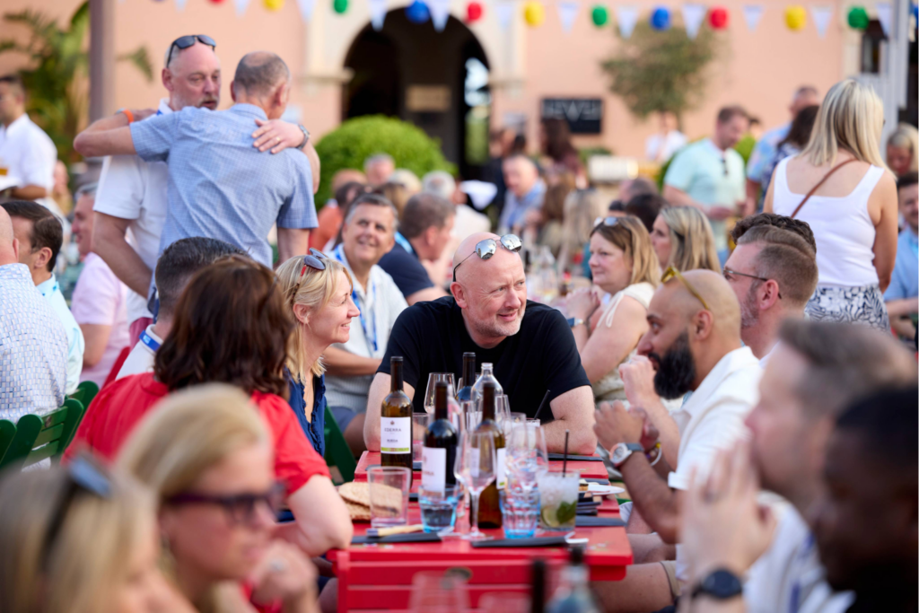 A large group of people sit at outdoor tables, eating, drinking, and socialising on a sunny day. Colourful flags and a bespoke exhibition stand add to the lively, cheerful scene.