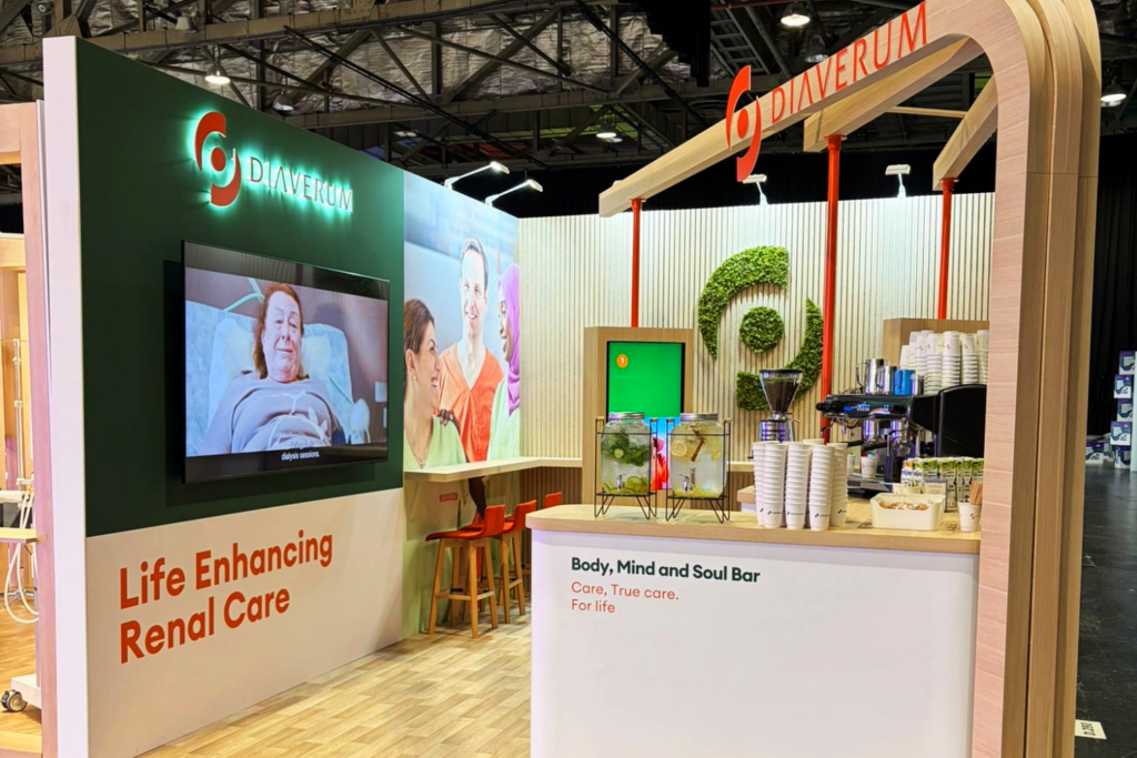 A modern bespoke exhibition stand for Diaverum features a screen showing a patient in bed, a coffee bar with cups and equipment, and signage reading Life Enhancing Renal Care and Body, Mind and Soul Bar.