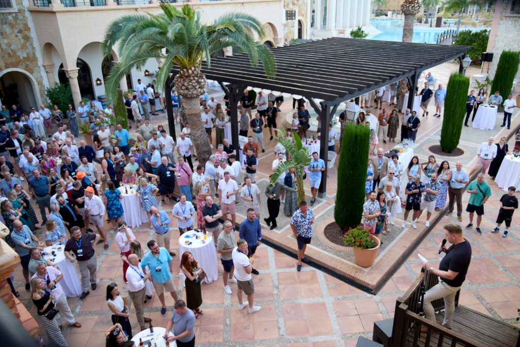 A large group of people gather in an outdoor courtyard, listening to a person speaking on a raised platform beside a bespoke exhibition stand. Tall palm trees, potted plants, cocktail tables, and Mediterranean-style architecture are visible.