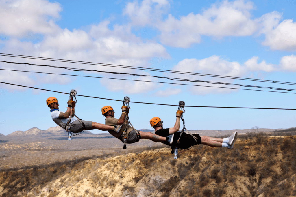 Three people wearing helmets and harnesses are ziplining together in a row, holding onto each other as they glide horizontally above a hilly landscape under a partly cloudy sky, enjoying thrilling shared experiences.