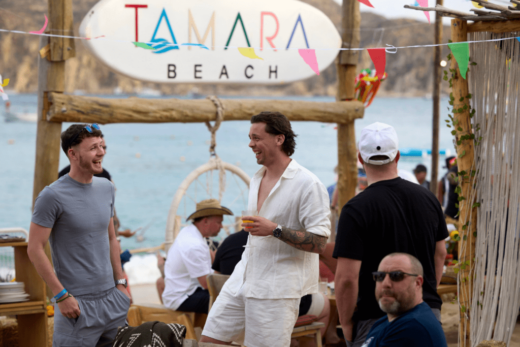 Several people stand and sit at the outdoor beach bar Tamara Beach, enjoying shared experiences. Two men chat and smile, one holding a drink, with festive decorations and the sea in the background.