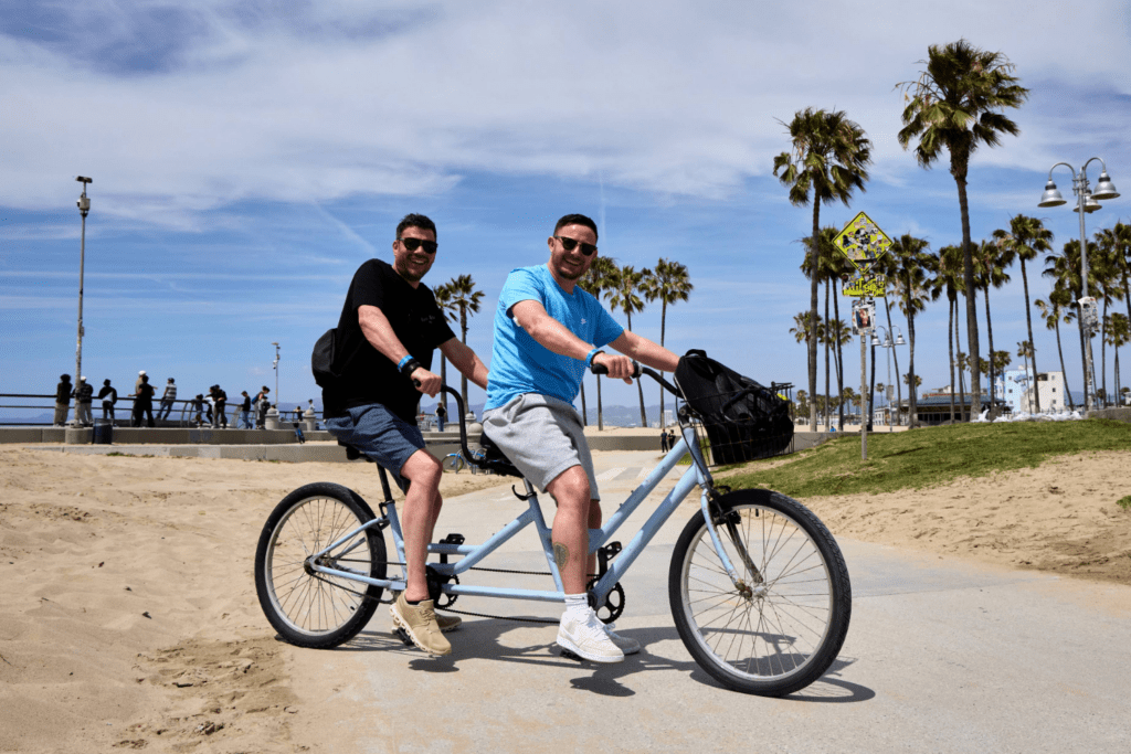 Two men enjoy shared experiences as they ride a tandem bicycle along a sunny beach path lined with palm trees. Both are smiling in casual summer clothes and sunglasses, soaking in the blue sky and warm atmosphere.