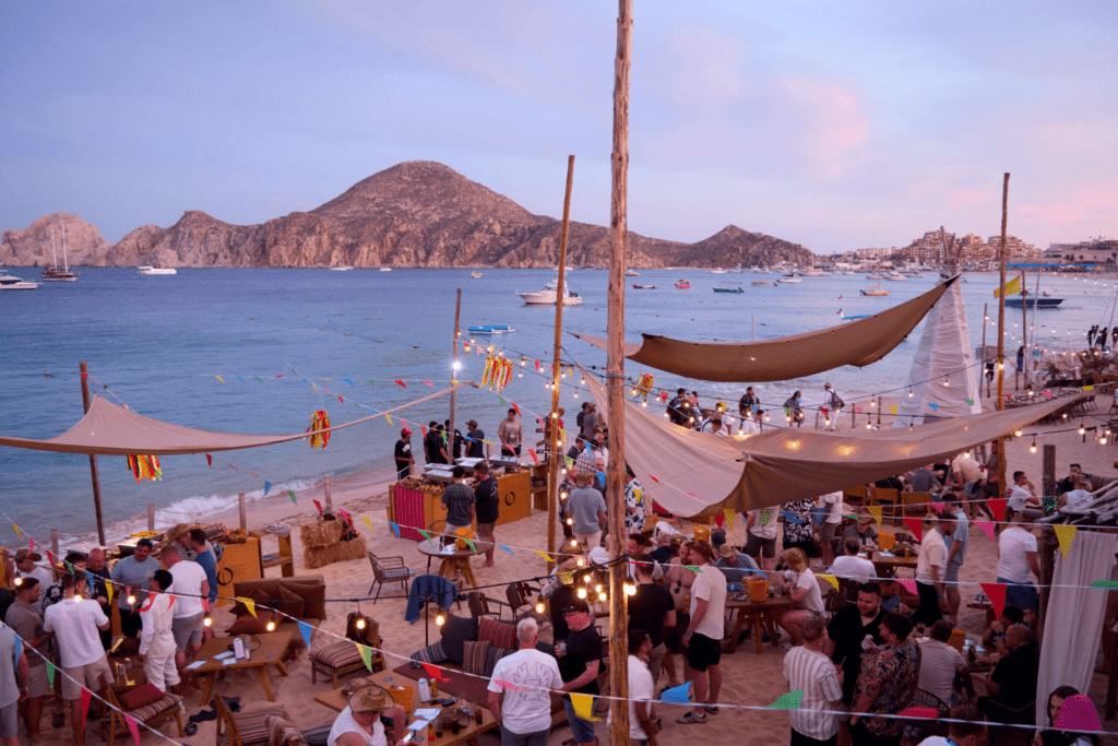 A lively beach party at sunset, where shared experiences bring crowds together under fairy lights and colourful flags. Tents offer shade, with mountains rising in the background near the calm sea dotted with boats.