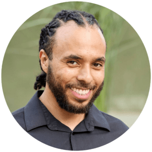A smiling man with short, plaited hair and a beard, wearing a black collared shirt, stands outdoors with greenery blurred in the background—looking ready to design your next Bespoke Exhibition Stand.