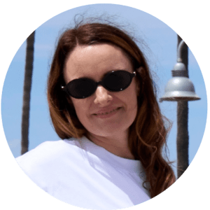 A woman with long brown hair wearing black oval sunglasses and a white shirt smiles outdoors, enjoying shared experiences among palm trees and a streetlamp under a blue sky.