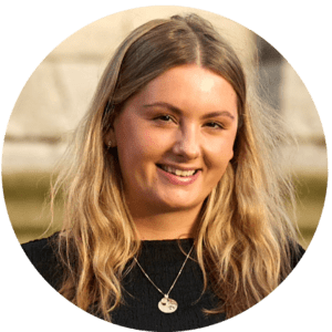 A young woman with long blonde hair, wearing a black top and a necklace with a round pendant, smiles at the camera, radiating warmth and the joy of shared experiences. The softly blurred outdoor background adds a gentle touch.