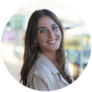 A woman with long brown hair and blue eyes smiles at the camera, her beige jacket and gold hoop earrings reflecting warmth. The blurred indoor background hints at shared experiences unfolding just beyond the frame.