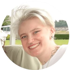 A person with short, platinum blonde hair and facial piercings smiles outdoors, their tattoos on display. Wearing a white shirt, they stand on a green lawn with trees behind them, radiating confidence and the warmth of shared experiences.