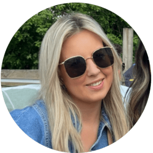 A woman with long blonde hair wearing large, square black sunglasses and a blue denim shirt smiles outdoors, embracing shared experiences amid vibrant greenery in the background.