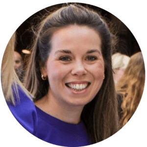 A woman with long brown hair, wearing a purple shirt and gold hoop earrings, smiles at the camera. The blurred background hints at shared experiences with the people standing behind her.