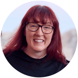 A woman with long red hair and a fringe, wearing glasses and a black top, smiles at the camera against a light, blurred outdoor background at an industry conference.