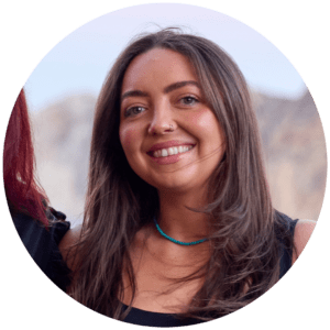 A woman with long brown hair, wearing a black top and a turquoise beaded necklace, smiles at the camera. The background is blurred, hinting at an outdoor setting during an industry conference.