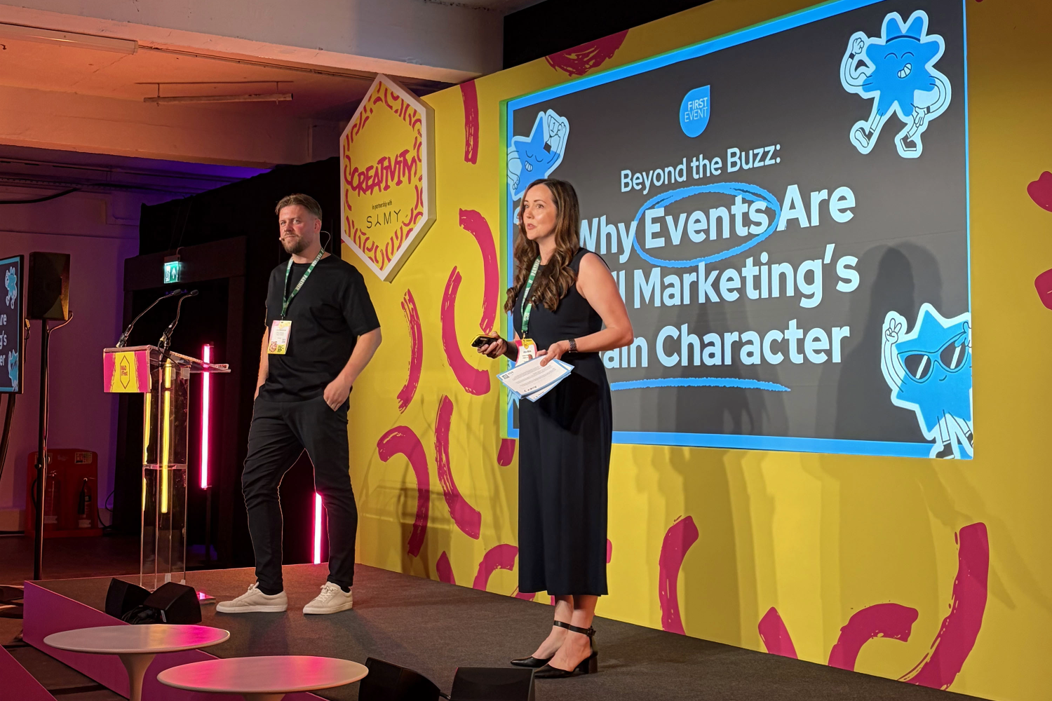 A man and woman present on stage at a marketing event, highlighting the importance of events in marketing. The screen behind them reads, “Why Events Are Marketing’s Main Character,” while bright, colourful signs and abstract designs decorate the area.