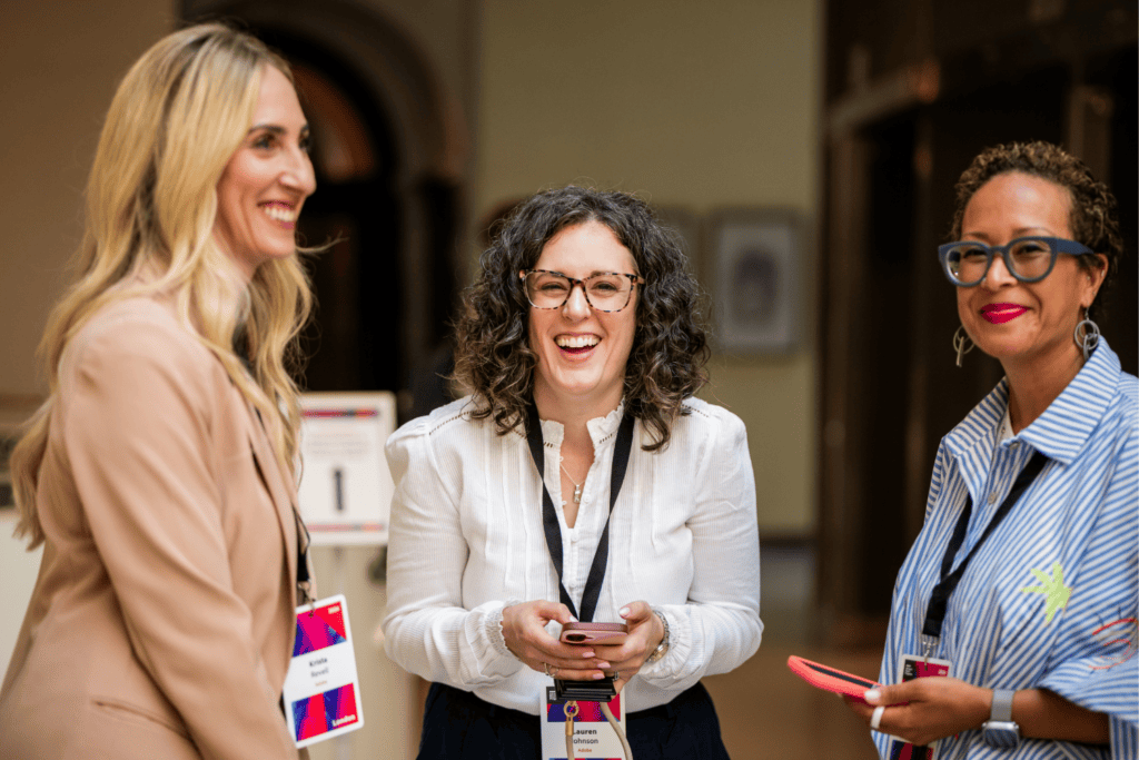 Three women with conference badges stand indoors at an advisory board event, smiling and talking. One holds a mobile. They appear happy and engaged in conversation. The background is softly blurred.