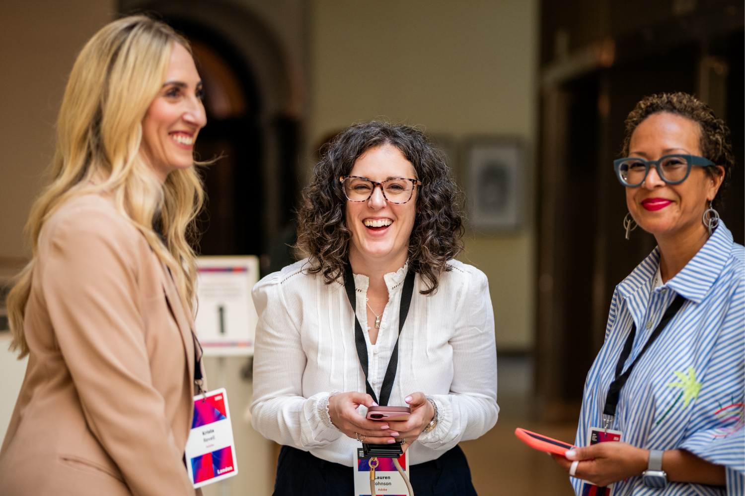 Three women with conference badges stand indoors at an advisory board event, smiling and talking. One holds a mobile. They appear happy and engaged in conversation. The background is softly blurred.