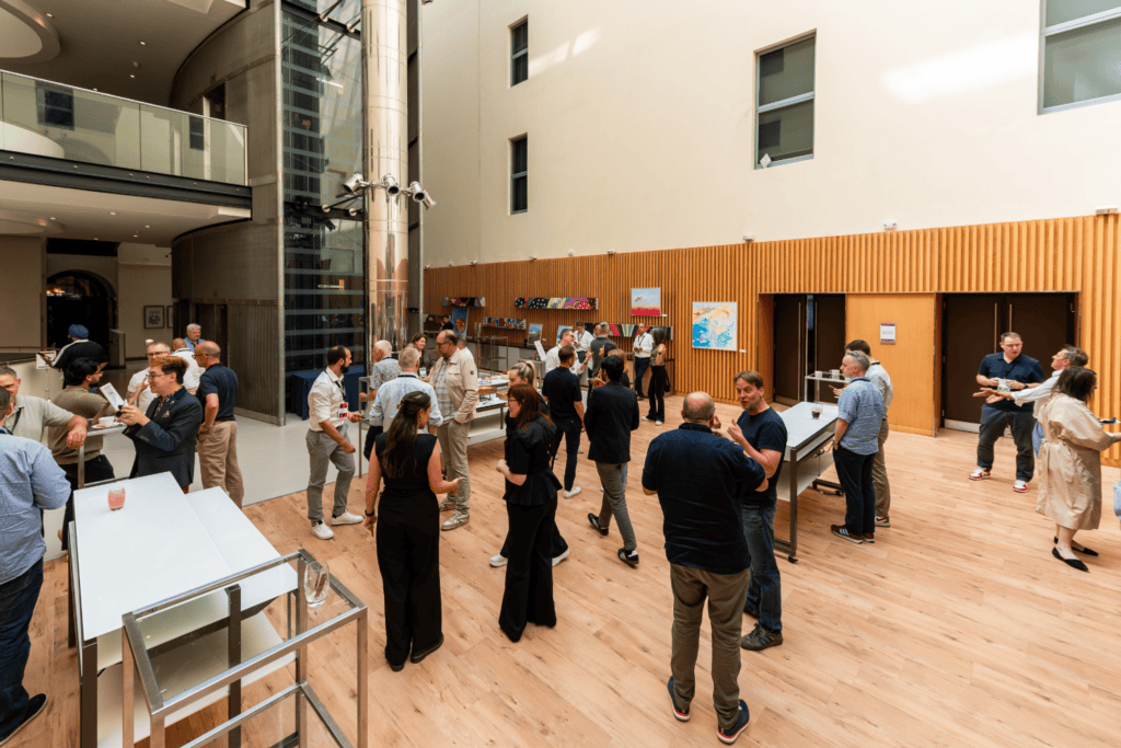 People mingle and converse in a spacious, modern indoor area with wooden flooring, high ceilings, artwork on the walls, and natural light coming through large windows during an engaging advisory board event.