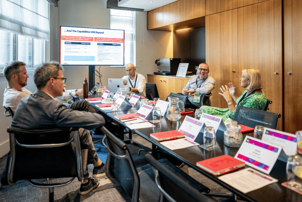 Six people sit around a conference table with documents and name cards, engaged in discussion during an advisory board event. A large monitor displays a presentation slide. The room is modern, with wooden cupboards and natural light from windows.