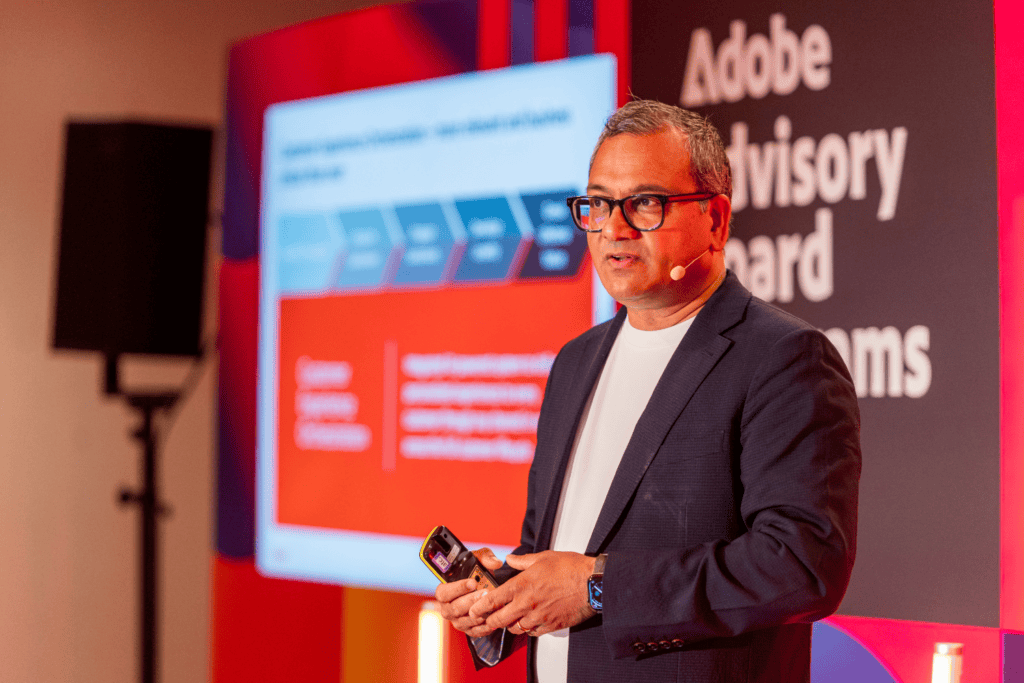 A man in a blazer and glasses speaks on stage at the Adobe Advisory Board event, holding a device. During this advisory board event, a presentation with a red and blue graphic is displayed behind him.