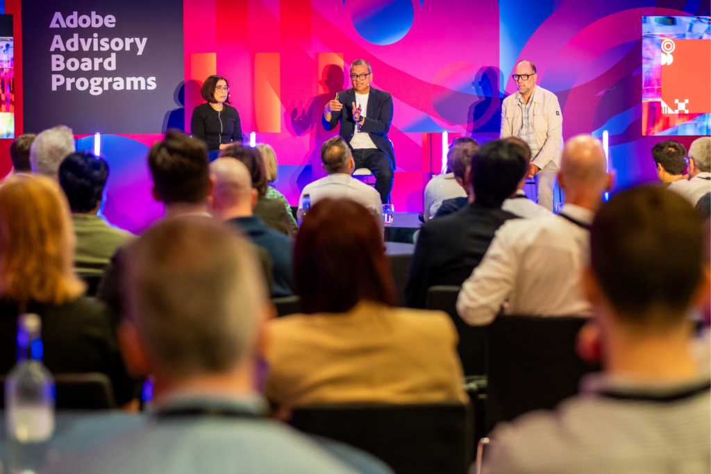A panel of three people speaks on stage at an Advisory Board event as part of the Adobe Advisory Board Programmes, facing an audience. The stage features colourful lighting and branding, with attendees seated and listening.