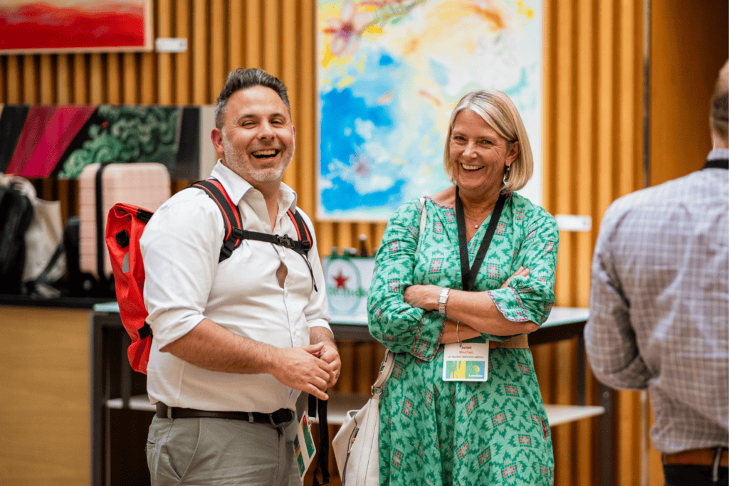 At an advisory board event, two people are smiling and talking indoors. The man wears a white shirt and red rucksack; the woman wears a green dress. Colourful art and drinks are visible in the background.