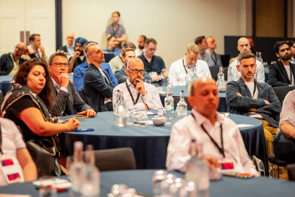 Attendees sit at round tables in a conference room, listening attentively to a speaker during the advisory board event. Bottles of water and glasses are on the tables. Most people appear engaged and focussed.
