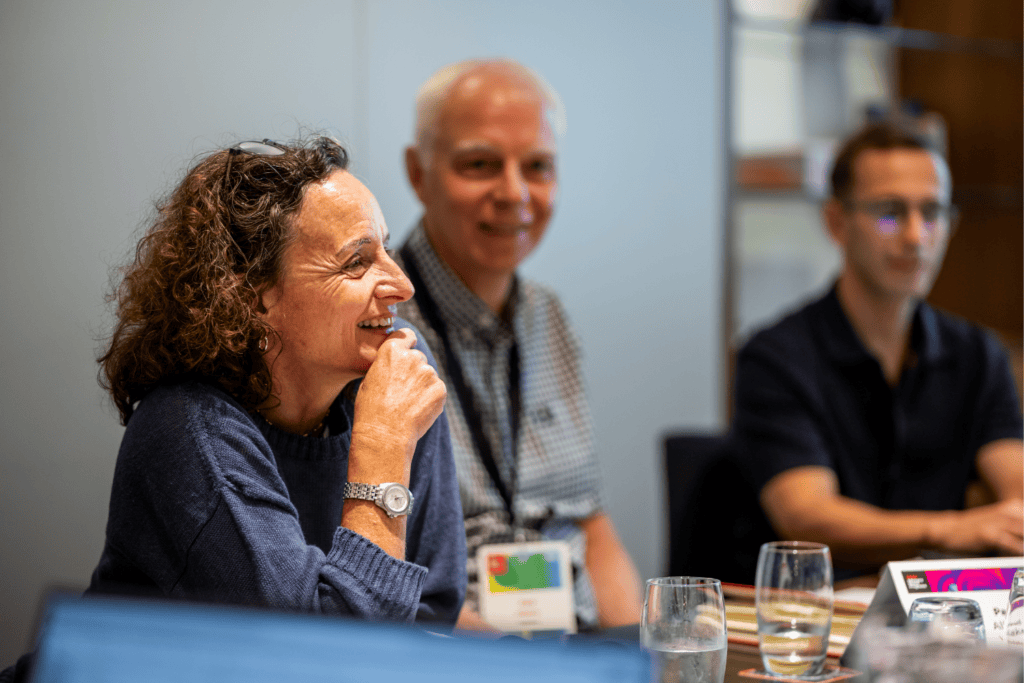 A woman smiles and listens attentively during an advisory board event, with two men seated beside her. There are glasses of water and name cards on the table. The background is softly focussed.