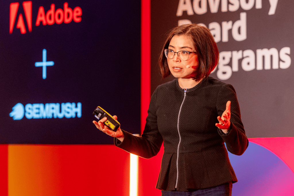 A woman wearing glasses speaks on stage with a microphone, holding a device during an advisory board event. Behind her are the logos of Adobe and SEMrush, along with partial text reading Advisory Board programmes. The lighting is bright and colourful.