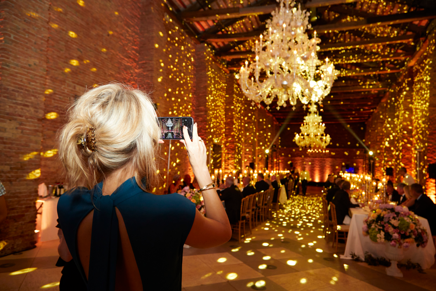 A woman with blonde hair takes a photo with her phone at an elegant, warmly lit indoor event with chandeliers and sparkling lights—a perfect scene for showcasing an effective event marketing strategy. Guests sit at decorated tables, enjoying the sophisticated atmosphere.