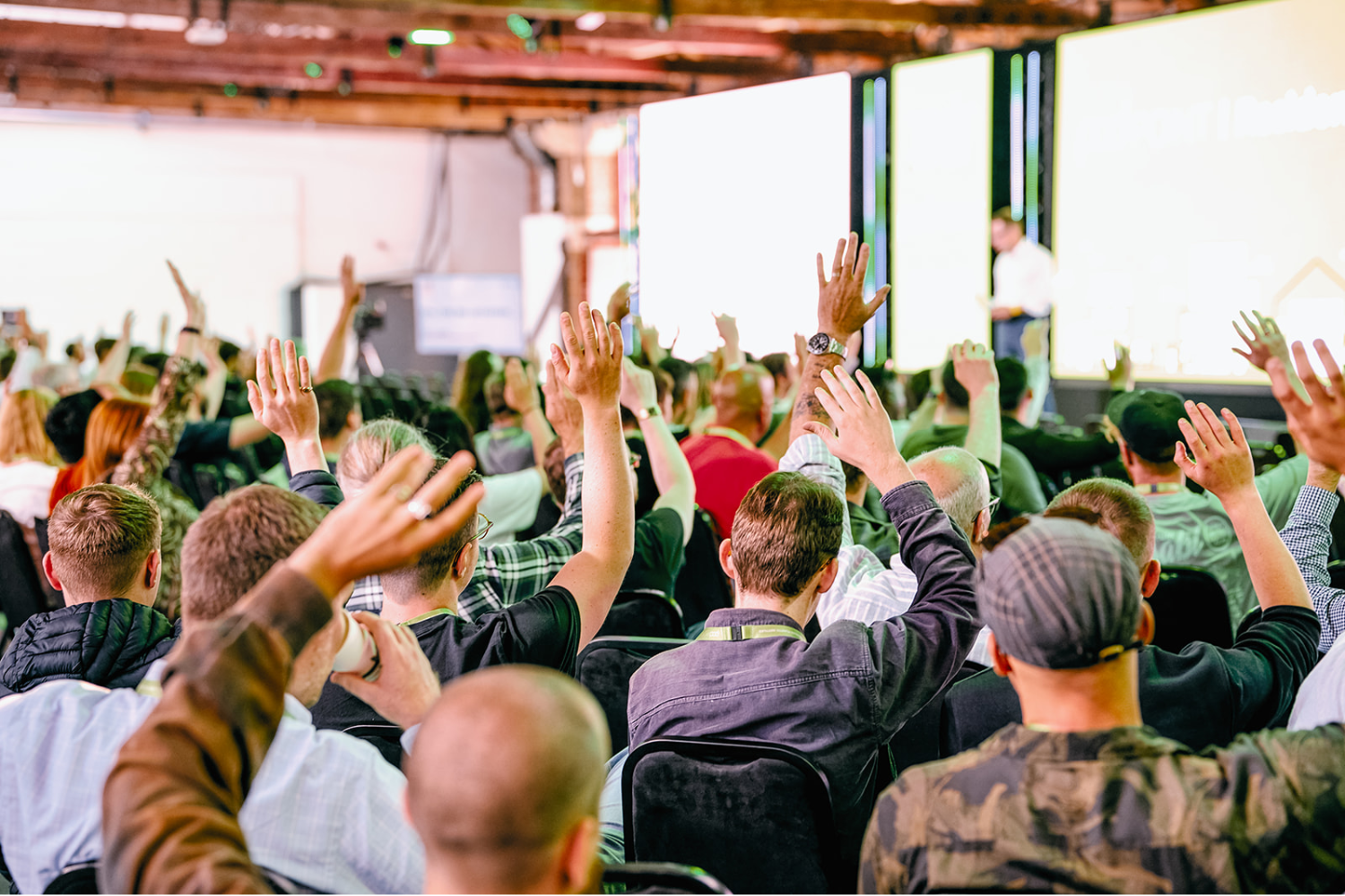 A group of people seated in an auditorium raise their hands towards a speaker presenting in front of large screens onstage in a brightly lit conference room, demonstrating high conference attendee engagement.