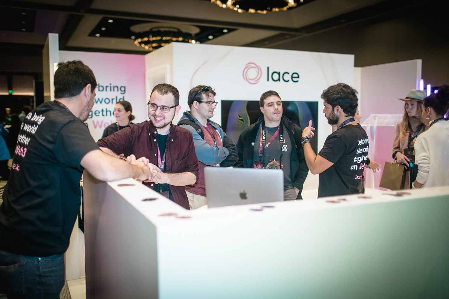 Several people stand and talk around a white stand with a lace logo at a conference or event, engaging in lively discussions about experiential marketing. Some wear conference badges and lanyards. A laptop is on the counter, and the atmosphere is friendly and dynamic.