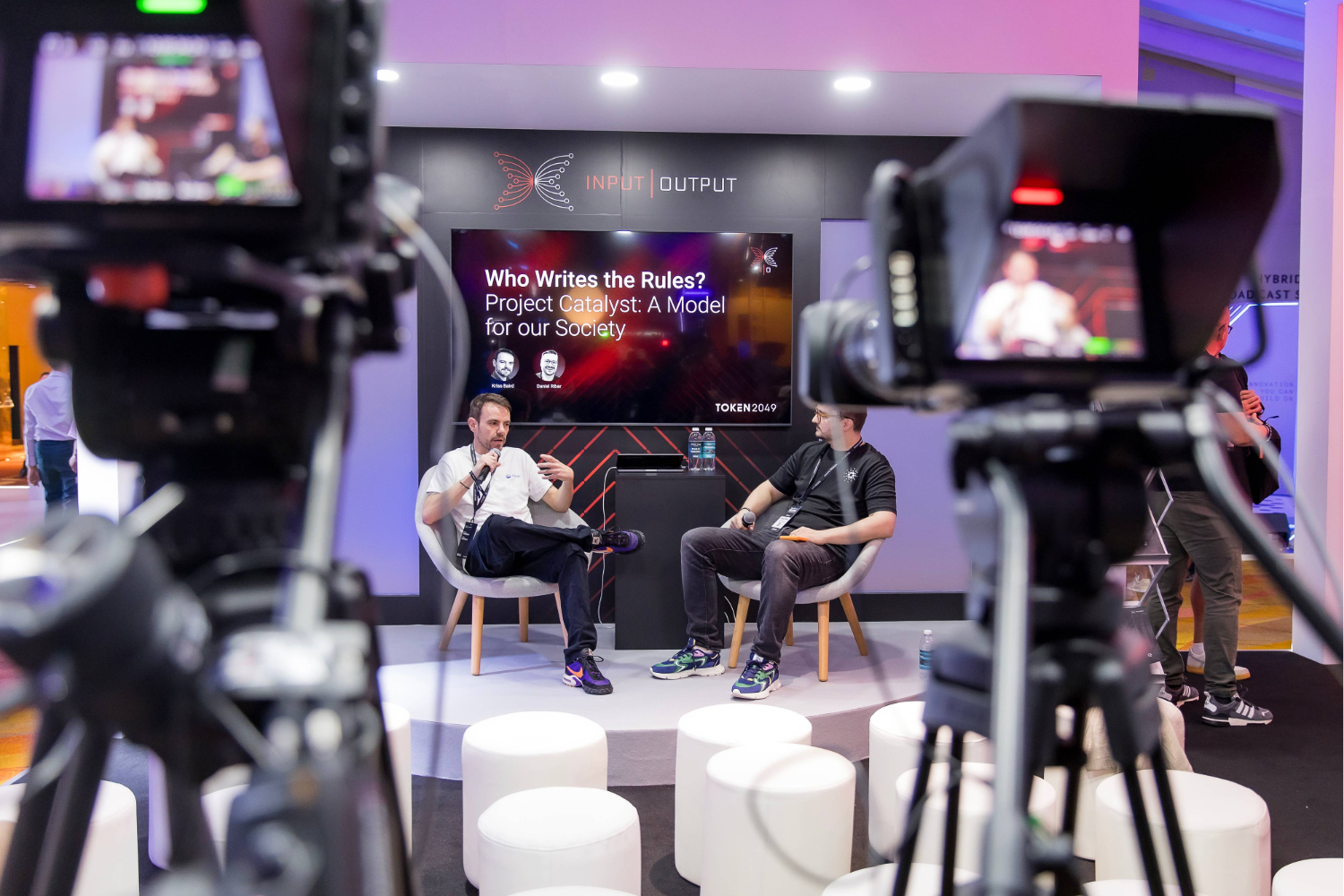 Two people sit on stage having a discussion, surrounded by cameras and lights—a clear example of hybrid business events in action. Behind them, a screen displays “Who Writes the Rules? Project Catalyst: A Model for our Society.”.
