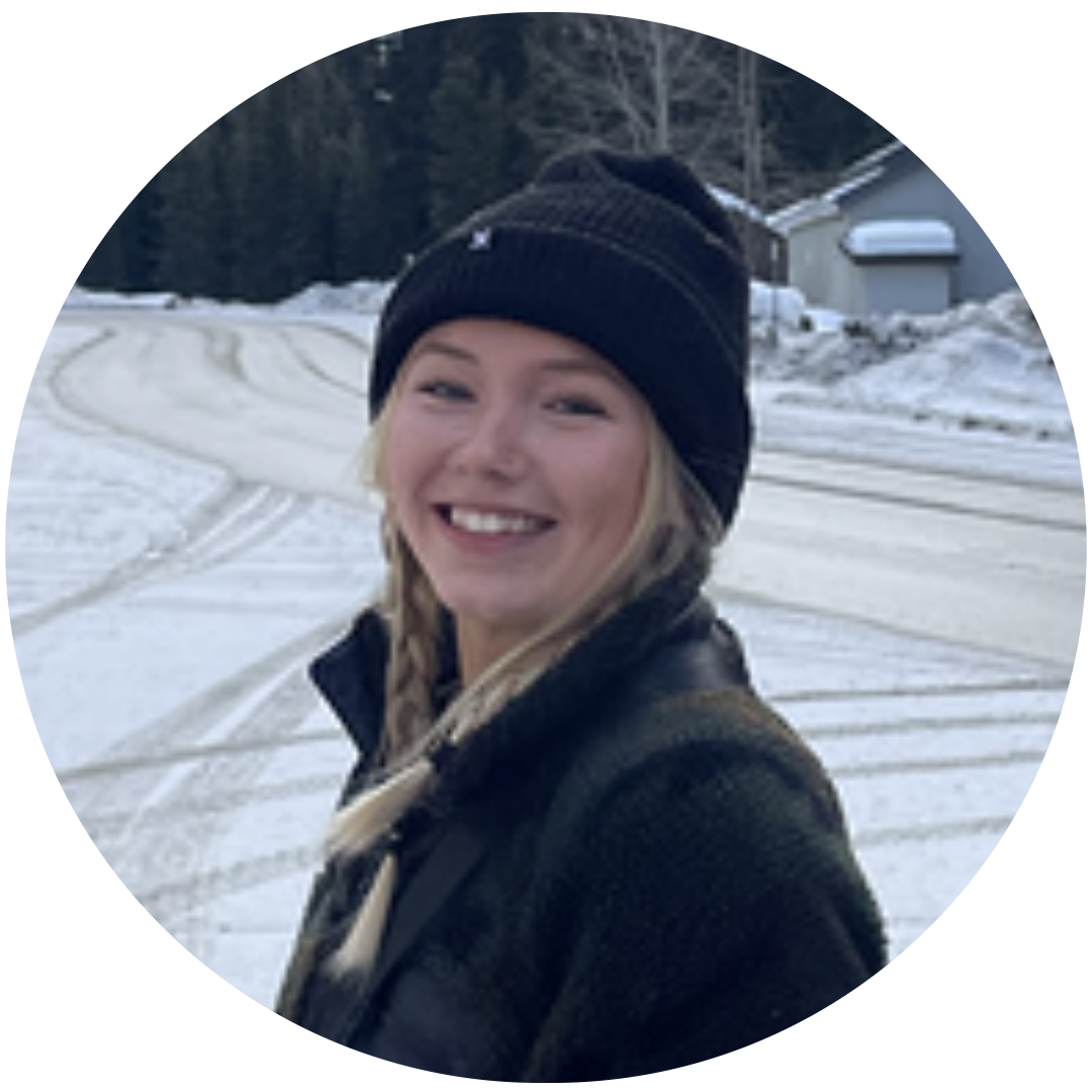 A young woman with blonde plaits, wearing a black beanie and dark jacket, smiles whilst standing outside on a snowy road with trees and houses in the background.