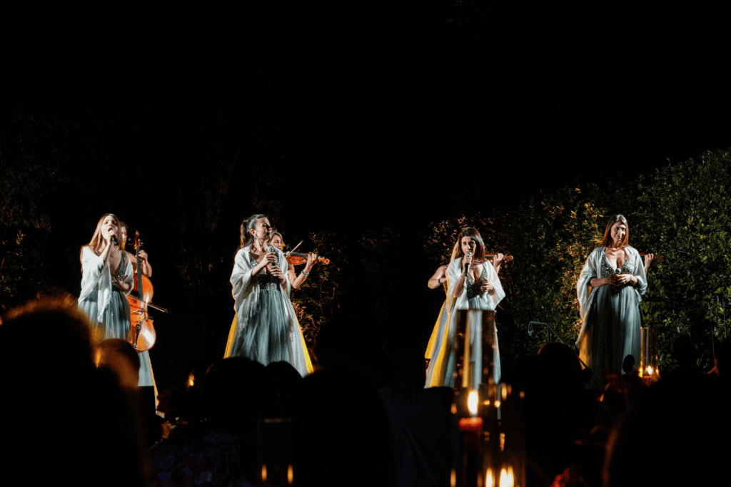 A group of women in light green dresses perform with string instruments and microphones on an outdoor, candle-lit stage at night, surrounded by greenery, creating a magical ambience perfect for an incentive travel programme.