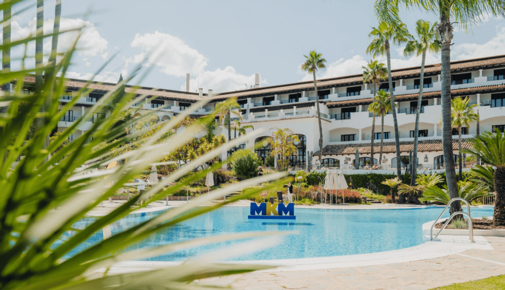A resort building with white walls and balconies overlooks a clear blue swimming pool, surrounded by palm trees and greenery. A large MKM sign floats in the pool—an inviting scene perfect for an incentive travel programme designed to reward and inspire achievement.