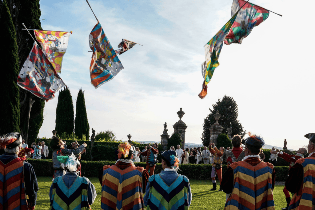 People in colourful, historical costumes watch performers toss vibrant flags into the air during an outdoor event surrounded by manicured gardens and tall trees—an unforgettable experience that could be a highlight of any incentive travel programme.