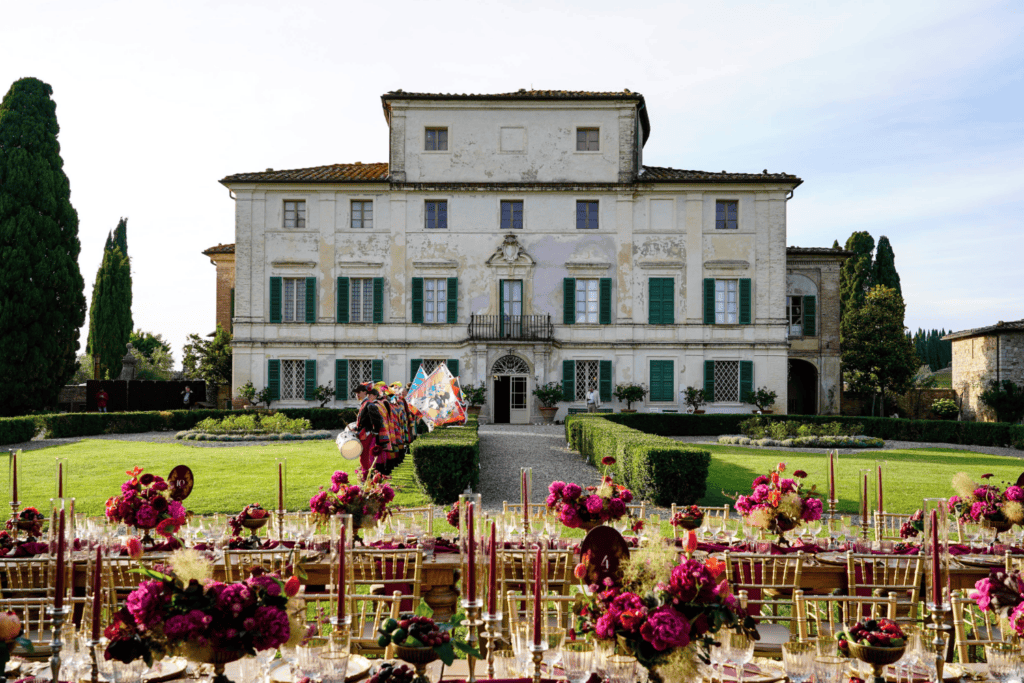 Long banquet tables with vibrant purple flowers and candles are set up in front of a large historic villa, creating a stunning setting for an incentive travel programme, as a group of people holding colourful flags walk along the gravel path towards the building.
